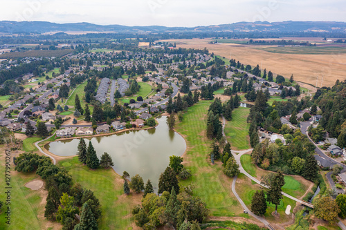 Aerial drone view of Staats Lake in Keizer, Oregon, near Salem, featuring a residential neighborhood, a golf course, and lush green landscapes under a clear blue sky