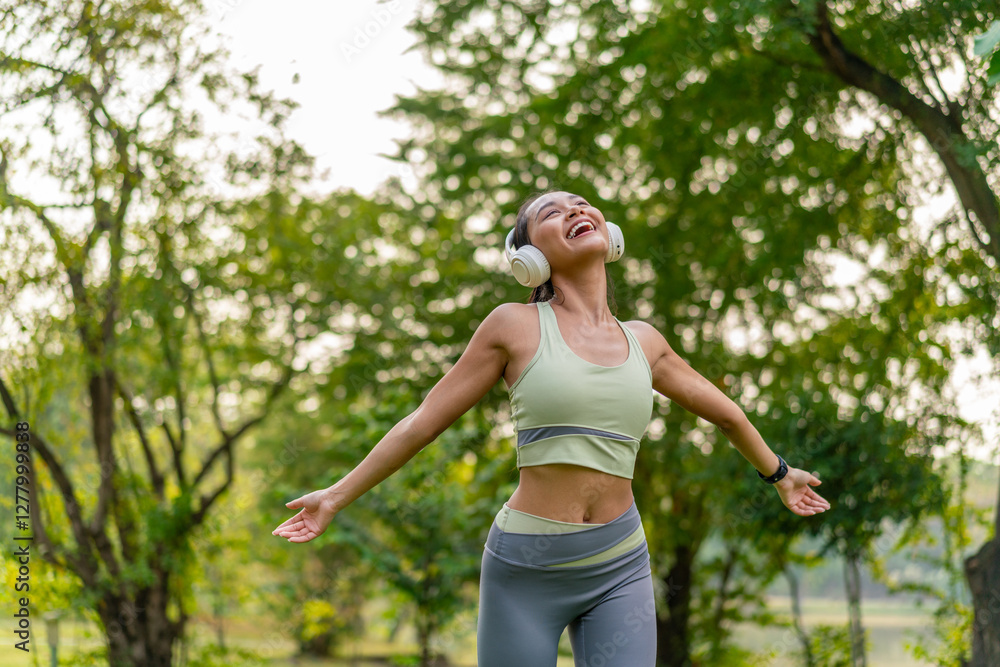 Young Asian woman enjoying music on headphones while exercising in green park. Sporty woman using headphone in outdoor nature setting. Young Asian woman enjoying music on headphones while exercising in green park. Sporty woman using headphone in outdoor nature setting.