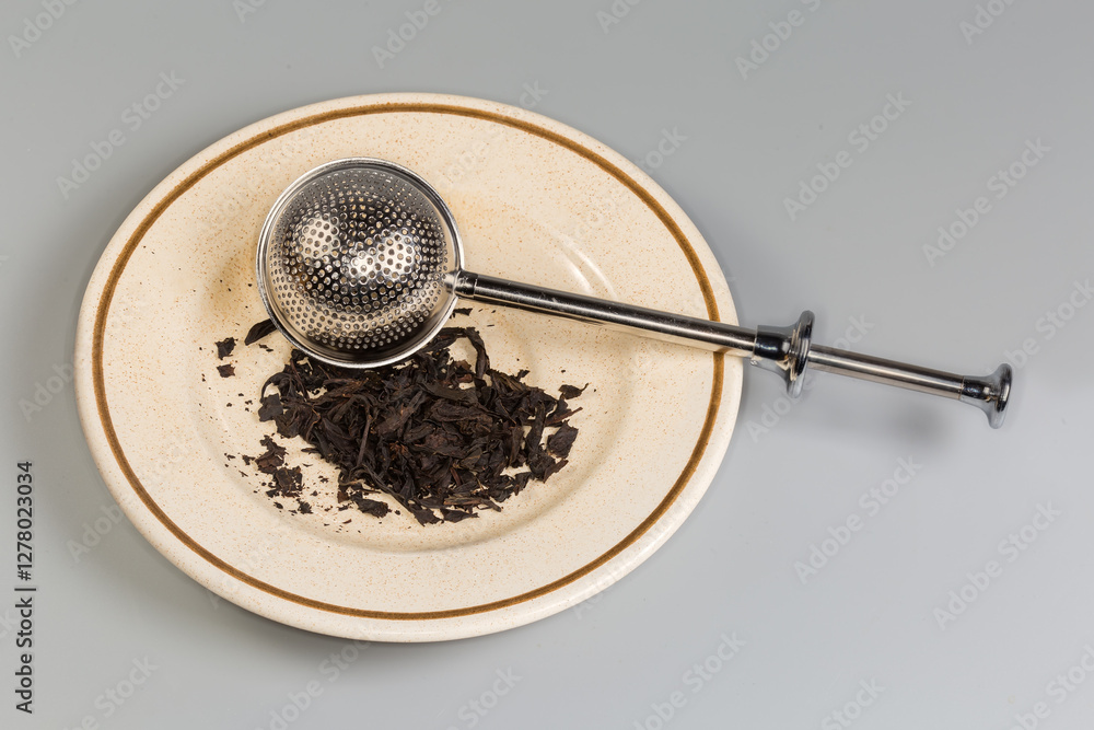 Fototapeta premium Stainless steel tea strainer on saucer with black tea leaves