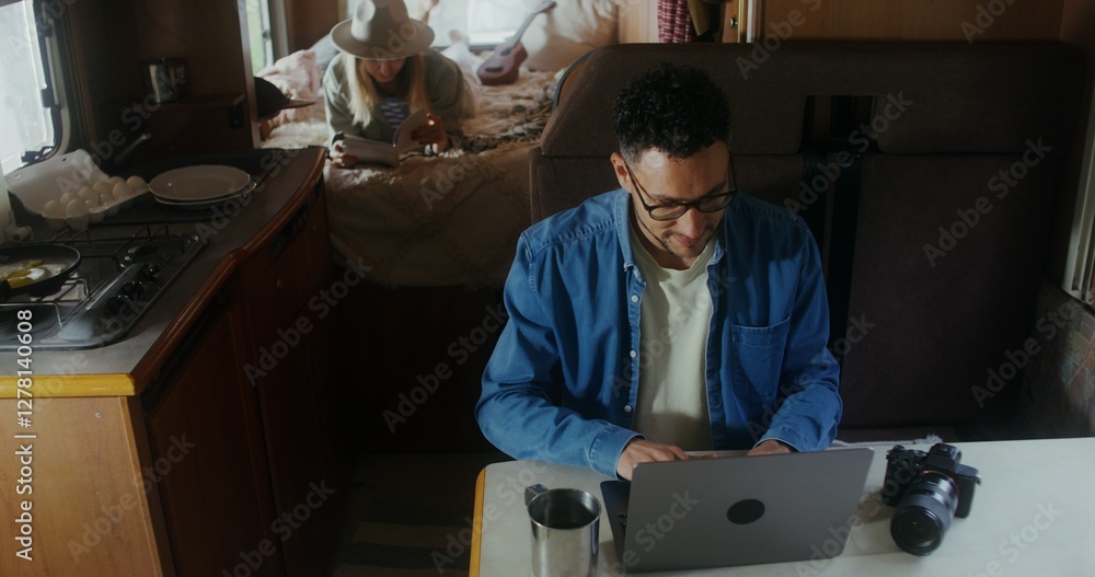 A young man uses a laptop and drinks a hot drink from a metal mug while sitting at a table in a van. A woman reads a book in the background A young man uses a laptop and drinks a hot drink from a metal mug while sitting at a table in a van. A woman reads a book in the background
