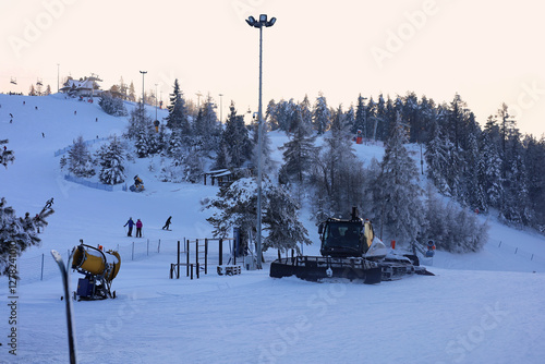 Fototapeta Naklejka Na Ścianę i Meble -  Snow cannon and parked snow groomer on the foreground. People having fun on the slope in the background