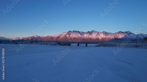 Unedited 4K aerial footage (59.94fps) of Knik River at sunset. Drone shots capture Knik River Bridge, Pioneer Peak, The Butte, and snow-capped mountains over frozen riverbed. photo