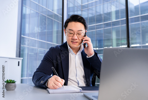 Confident businessman in formal attire discussing over phone call while taking notes in a notepad. Situated in a bright, glass-walled modern office space enhanced with natural light.