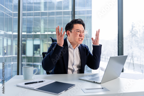 Upset Asian businessman expressing frustration at work in a professional environment. Modern office backdrop with natural lighting. Holding hands up while seated at desk using a laptop, coffee cup.
