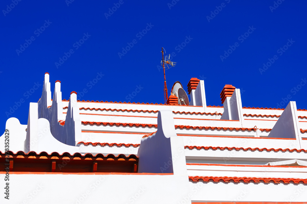 Tiered white building with terracotta colored roofing, showcasing architectural detail against a vibrant blue sky. White buildings with red-tiled roofs with multiple levels, chimneys, and an antenna