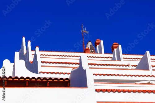 Obraz na płótnie Tiered white building with terracotta colored roofing, showcasing architectural detail against a vibrant blue sky. White buildings with red-tiled roofs with multiple levels, chimneys, and an antenna w salonie