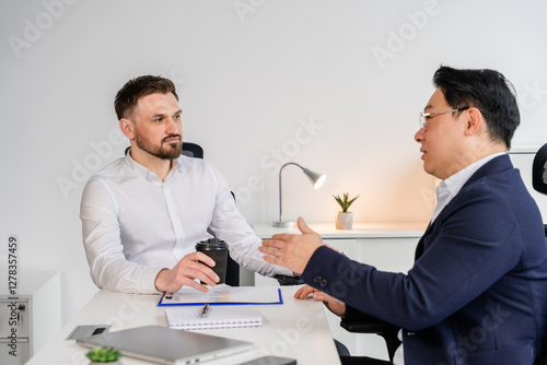 Modern business discussion between two professionally dressed individuals in a contemporary office environment, expressing focus and collaboration while sitting at a bright workstation.