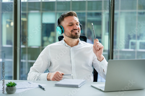 Professional businessman listening to music at a modern office desk, wearing headphones and enjoying a productive work environment. Surrounded by office tools and natural light from large windows.
