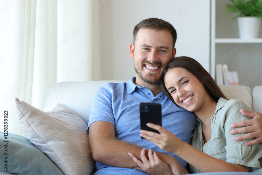 Happy couple holding phone looking at you at home Happy couple holding phone looking at you at home