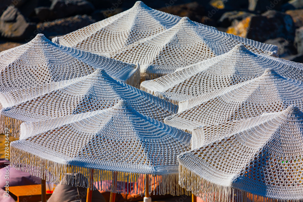 Woven white parasols arranged closely together, providing shade at beach. Detailed pattern of parasols fabric, along with fringe hanging from edges, creates an aesthetically pleasing