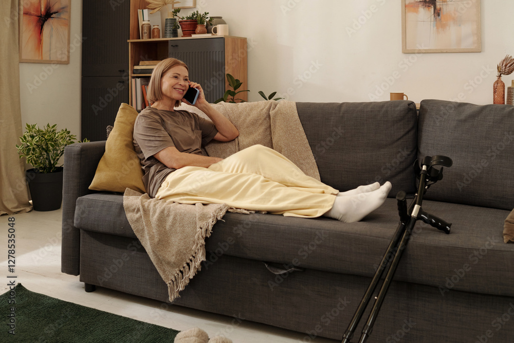 Obraz premium Woman sitting on couch while talking on phone and smiling warmly. Sofa with pillows, nearby shelves with books and plant add homely touch