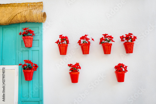 Vibrant red flower pots with blooming plants decorate a white wall beside turquoise window, creating striking contrast. Charming Mediterranean style exterior with fresh, colorful, and artistic design