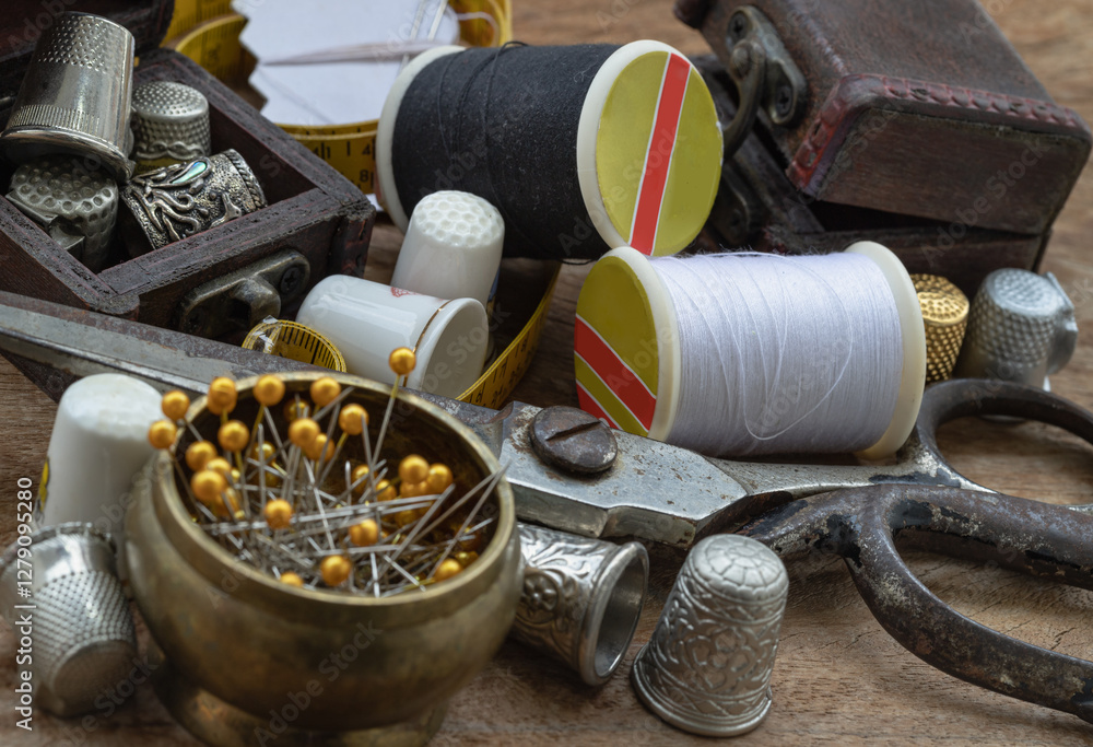 Sewing needles stuck in a white paper, Thimbles in small wooden treasure chest, Spools of black and white thread, Steel scissors and Measuring tape including pins in brass cup on wooden table. Sewing.