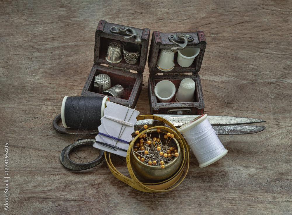 Thimbles in small wooden treasure chest, Sewing needles stuck in a white paper, Spools of black and white thread, Steel scissors and Measuring tape including pins in brass cup on wooden background.