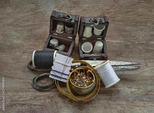 Obraz na płótnie Thimbles in small wooden treasure chest, Sewing needles stuck in a white paper, Spools of black and white thread, Steel scissors and Measuring tape including pins in brass cup on wooden background. w salonie