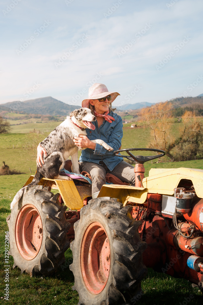 Obraz premium Smiling farmer woman with sunglasses and straw hat is sitting on tractor with her dog, enjoying a sunny day in the countryside