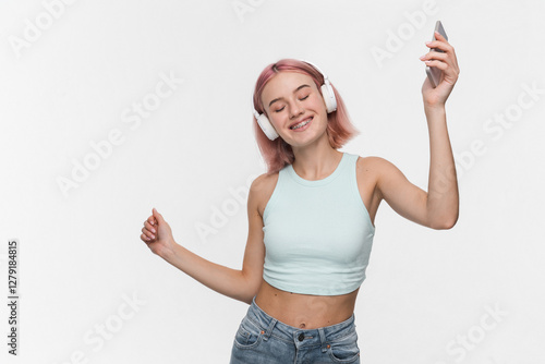 Positive stylish young girl female student wearing denim crop top standing isolated over white background, dancing listening to music song audio with wireless headphones holding smartphone.