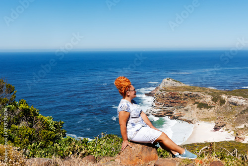 A slender woman with bright red hair sits on the rocky shore of Cape Agulhas, where the Indian and Atlantic Oceans meet, a cape in South Africa, the southernmost point of Africa.