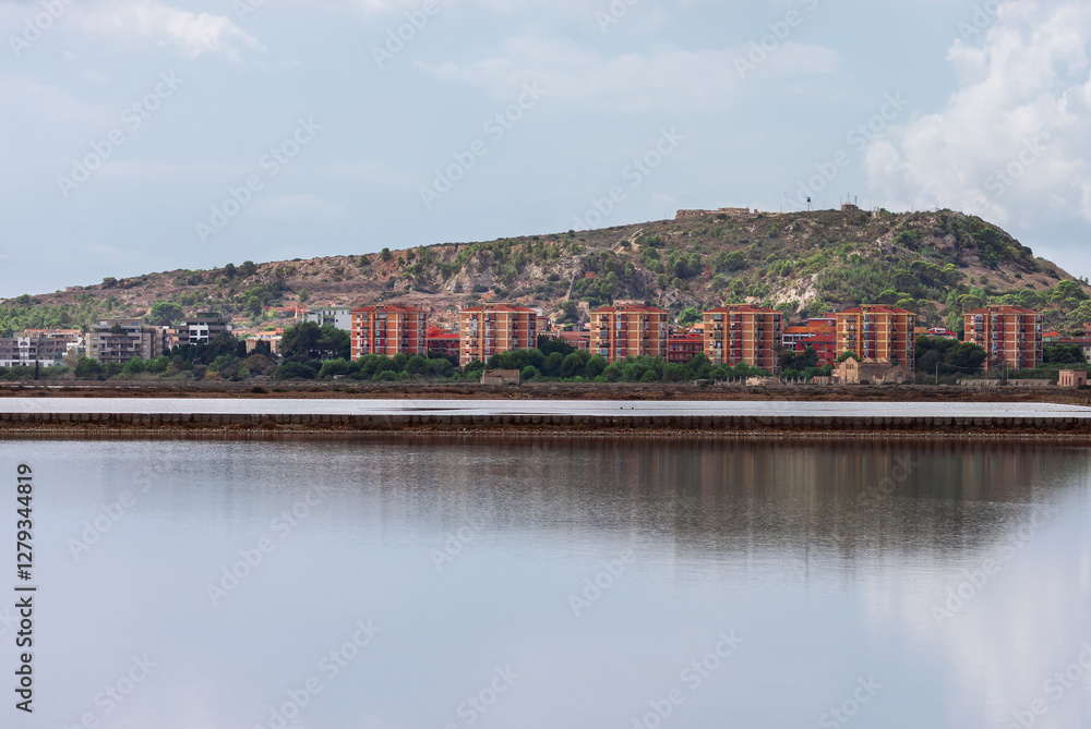Scenic view of Cagliari, Italy, featuring a calm body of water reflecting the cityscape and a hill in background. Cloudy sky adds depth to the serene atmosphere of this coastal urban landscape