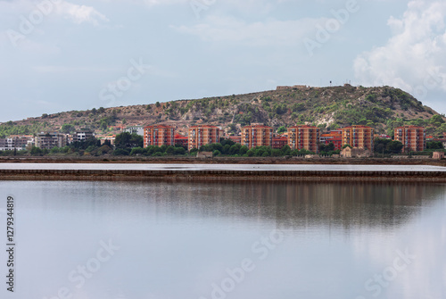 Obraz na płótnie Scenic view of Cagliari, Italy, featuring a calm body of water reflecting the cityscape and a hill in background. Cloudy sky adds depth to the serene atmosphere of this coastal urban landscape w salonie