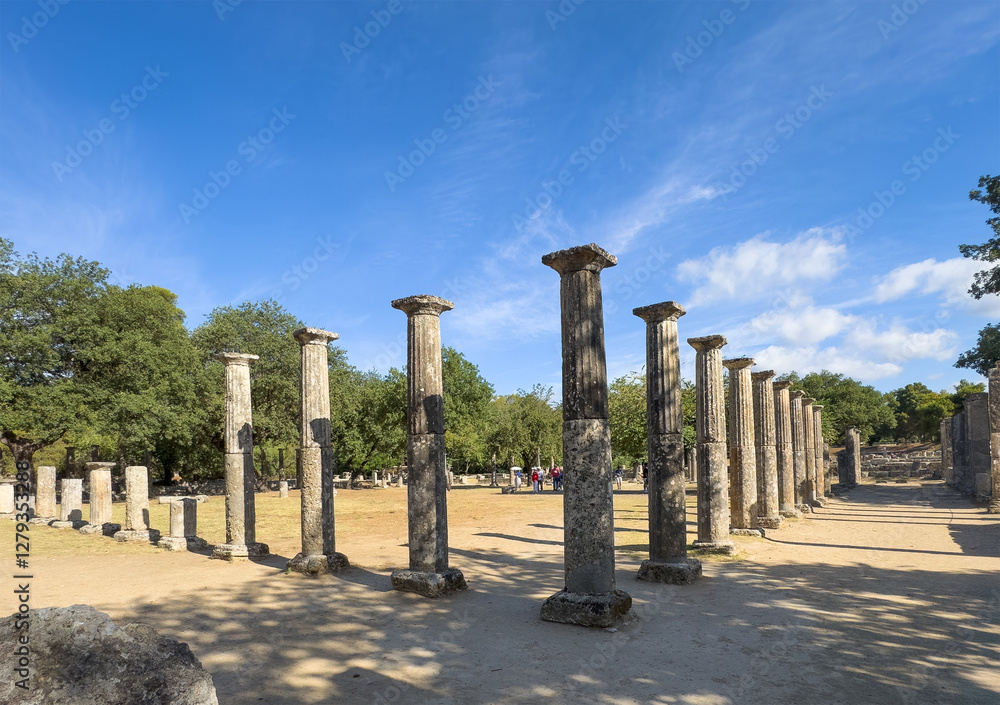 Olympia, Greece - July 4, 2024: Birthplace of the Olympic Games. South of Ancient Gymnasium. Rows of pillars. Green foliage belt behind and blue sky Olympia, Greece - July 4, 2024: Birthplace of the Olympic Games. South of Ancient Gymnasium. Rows of pillars. Green foliage belt behind and blue sky