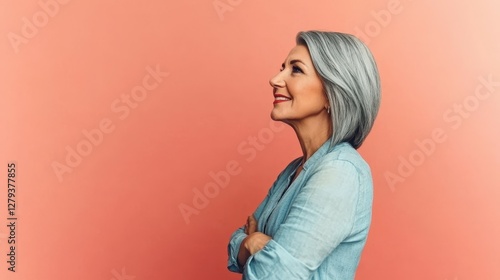 A confident older woman with silver hair smiles warmly while posing against a bright coral wall. She stands with her arms crossed, showcasing elegance and grace