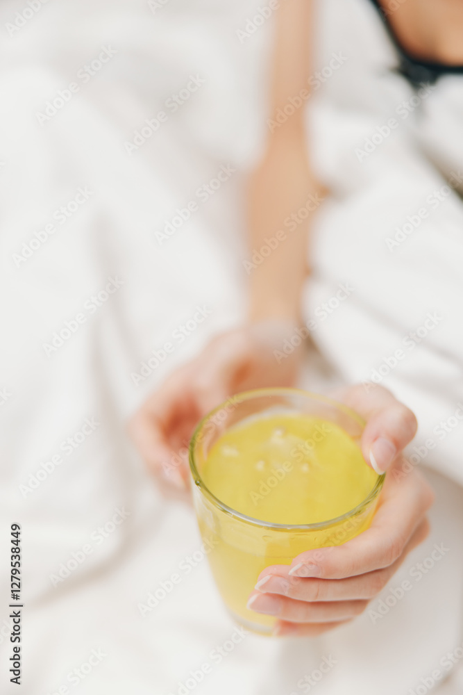Woman lying on a bed with a white sheet, holding a cup of yellow liquid in her hand Woman lying on a bed with a white sheet, holding a cup of yellow liquid in her hand