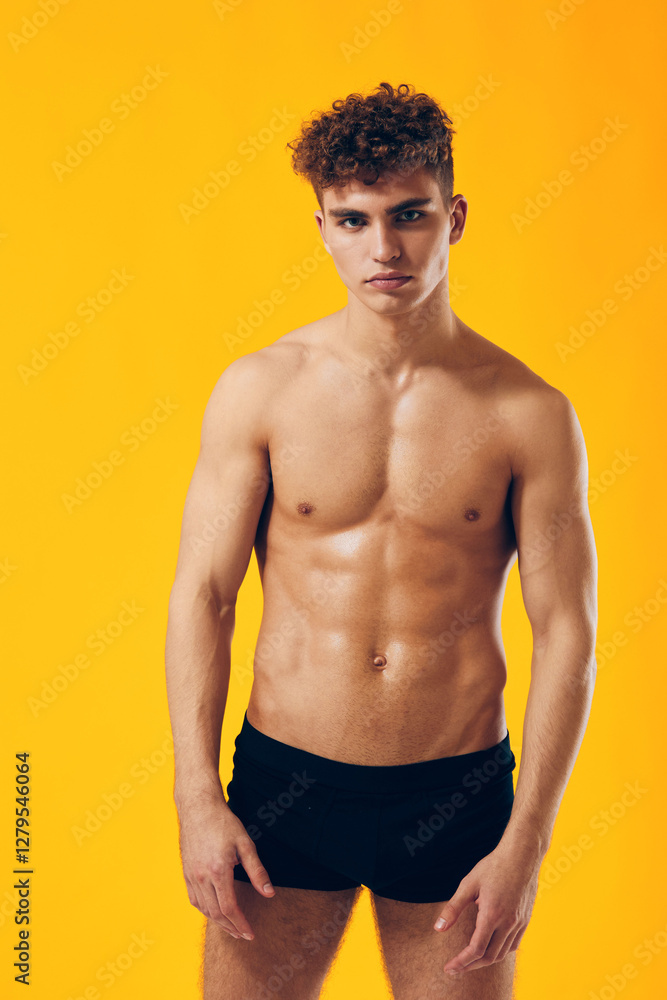 Fototapeta premium Confident young man with curly hair posing shirtless against a bright yellow background. Fitness and body positivity concept.
