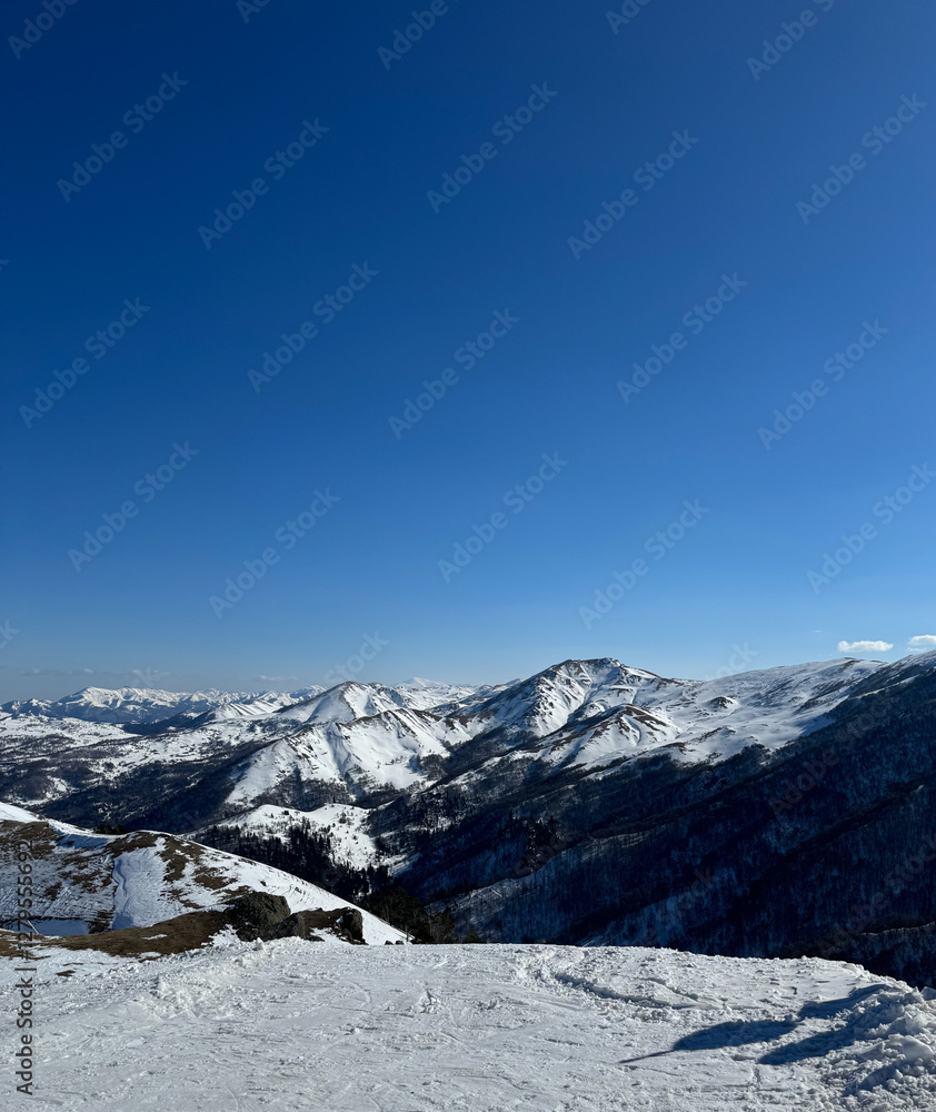 Fototapeta premium Vertical aerial view of the snow-covered mountain peaks in Bakuriani, Georgia, clear blue sky, with space for text, for cards and banner
