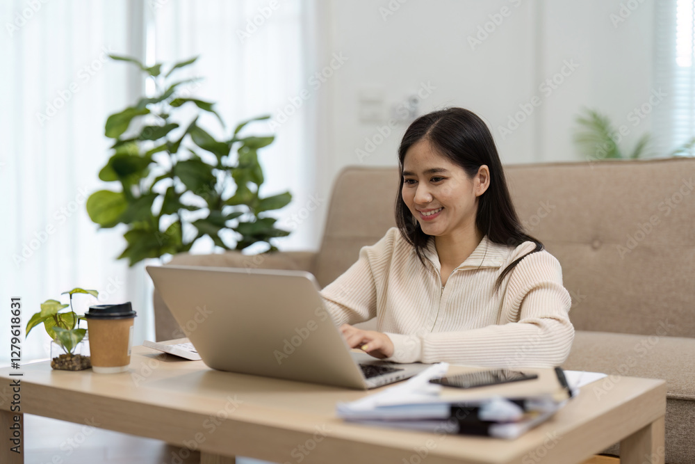 Young Asian woman smiling while working on a laptop at home, enjoying a productive and positive atmosphere. Young Asian woman smiling while working on a laptop at home, enjoying a productive and positive atmosphere.