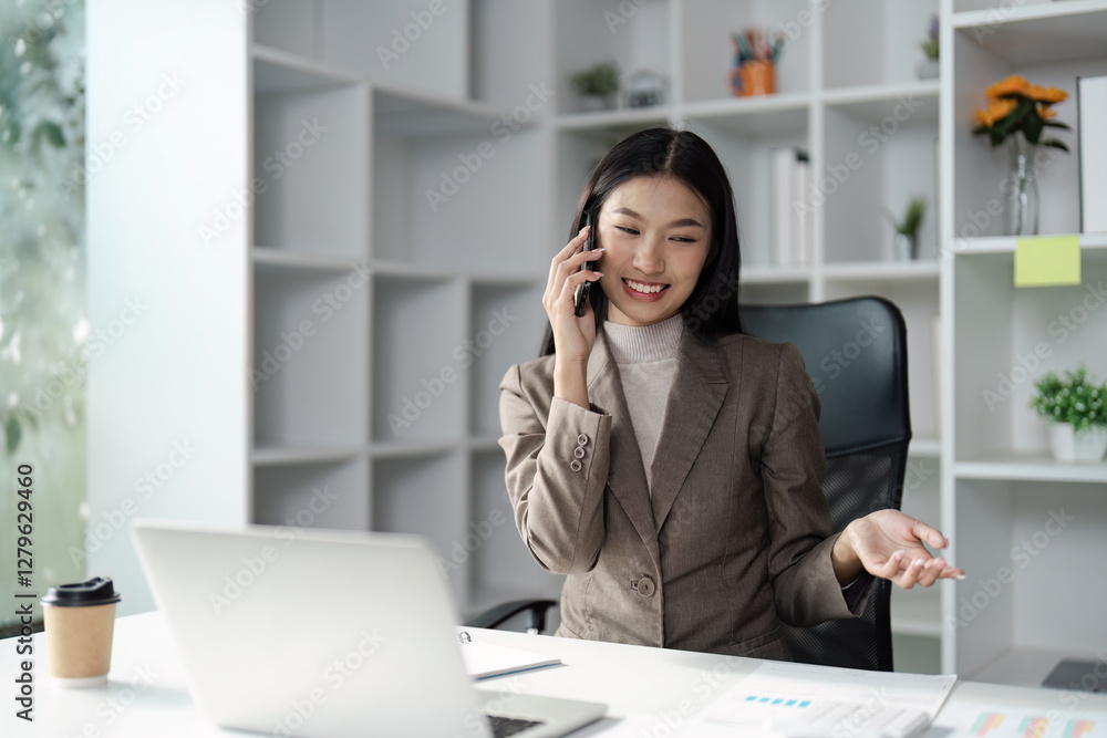 Young Asian businesswoman talking on the phone while working at a desk with a laptop in a modern office. Young Asian businesswoman talking on the phone while working at a desk with a laptop in a modern office.