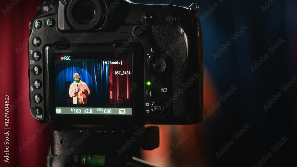 Closeup shot of mini digital camera screen on tripod filming Black male stand-up comedian performing on small stage with red and blue curtains Closeup shot of mini digital camera screen on tripod filming Black male stand-up comedian performing on small stage with red and blue curtains