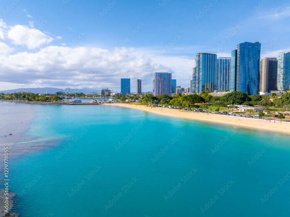 Aerial view of Honolulu, Oahu, Hawaii, showing turquoise waters, sandy beach, greenery, a road with vehicles, and modern high rise buildings under a blue sky. Aerial view of Honolulu, Oahu, Hawaii, showing turquoise waters, sandy beach, greenery, a road with vehicles, and modern high rise buildings under a blue sky.