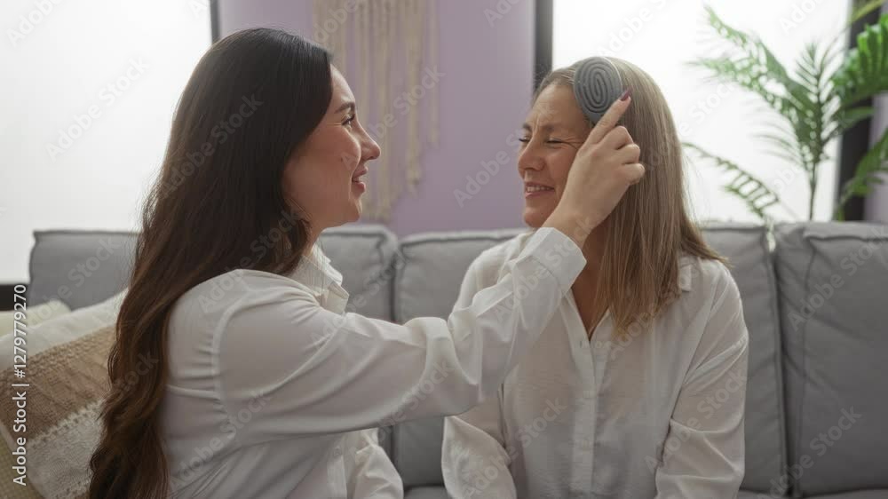 Mother and daughter share a loving moment indoors as the daughter gently combs her mother's hair in a cozy living room setting, showcasing family bond and warmth. Mother and daughter share a loving moment indoors as the daughter gently combs her mother's hair in a cozy living room setting, showcasing family bond and warmth.