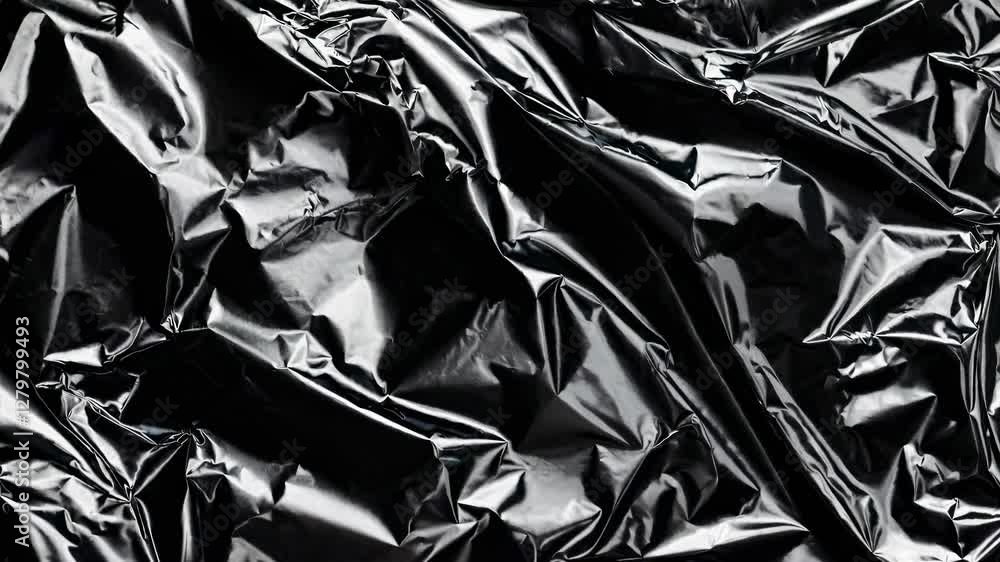 Close-up shot of crumpled black foil, creating an abstract, textured background. The video captures the shiny, reflective surface from a top-down angle. Close-up shot of crumpled black foil, creating an abstract, textured background. The video captures the shiny, reflective surface from a top-down angle.