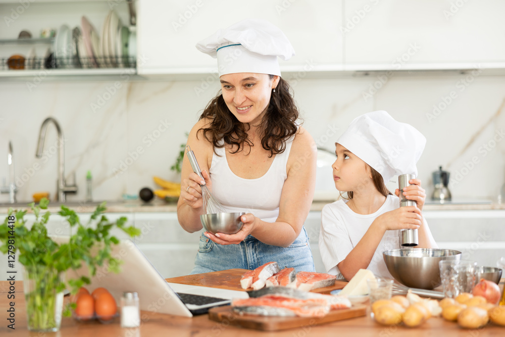 Happy mother and daughter are baking salmon in the kitchen while looking at the recipe on laptop Happy mother and daughter are baking salmon in the kitchen while looking at the recipe on laptop