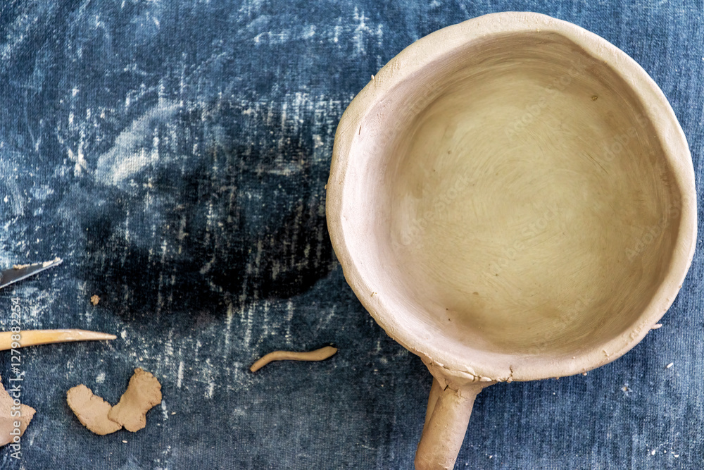 Fototapeta premium Handcrafted, unfired clay bowl with a handle sits on a textured blue surface. Surrounding the bowl are small pieces of clay and sculpting tools