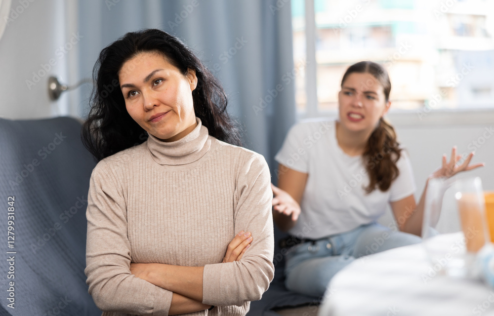 Upset young woman sitting at home table with disgruntled woman behind Upset young woman sitting at home table with disgruntled woman behind