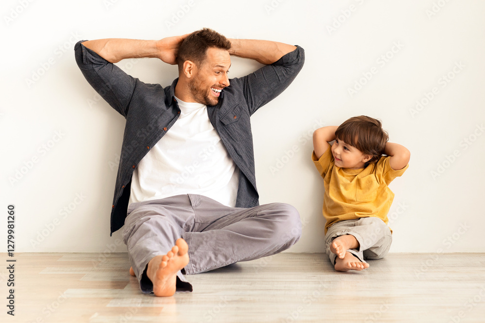 Common family pastime. Happy young man smiling to his cute little son, relaxing together at home, leaning wall with hands behind heads, white background, full length Common family pastime. Happy young man smiling to his cute little son, relaxing together at home, leaning wall with hands behind heads, white background, full length