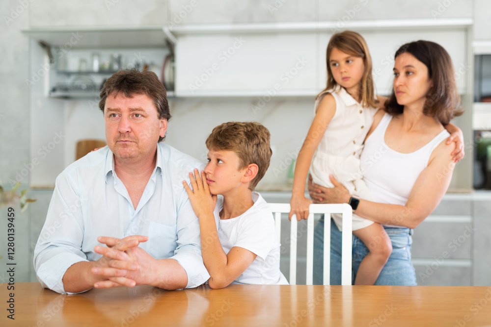 Offended father sitting at the kitchen table being hugged by little son, his wife and daughter looking at them with empathy Offended father sitting at the kitchen table being hugged by little son, his wife and daughter looking at them with empathy