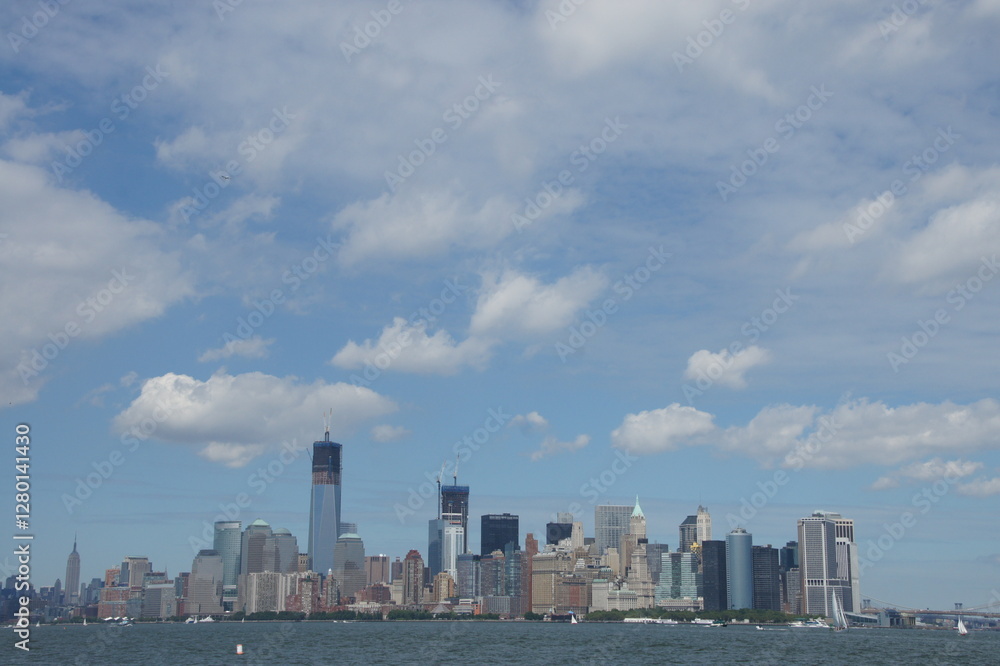 Fototapeta premium New York, Manhattan boasts a towering skyline with the Statue of Liberty standing proudly in the foreground. The calm water reflects the tall skyscrapers against a clear blue sky, creating an iconic