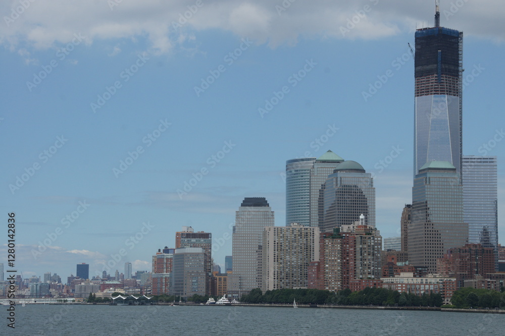 Obraz premium A vibrant New York, Manhattan skyline bathed in the warm glow of a sunset, with skyscrapers casting their reflections onto the still waters of the Hudson River. Bustling city life continues along