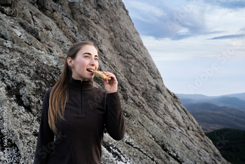 Friendly woman hiker eating granola muesli bar standing in the mountains. Female traveler eating a grain cereal snack on the hike, woman takes a break with dessert. Healthy nutrition, tourism. photo