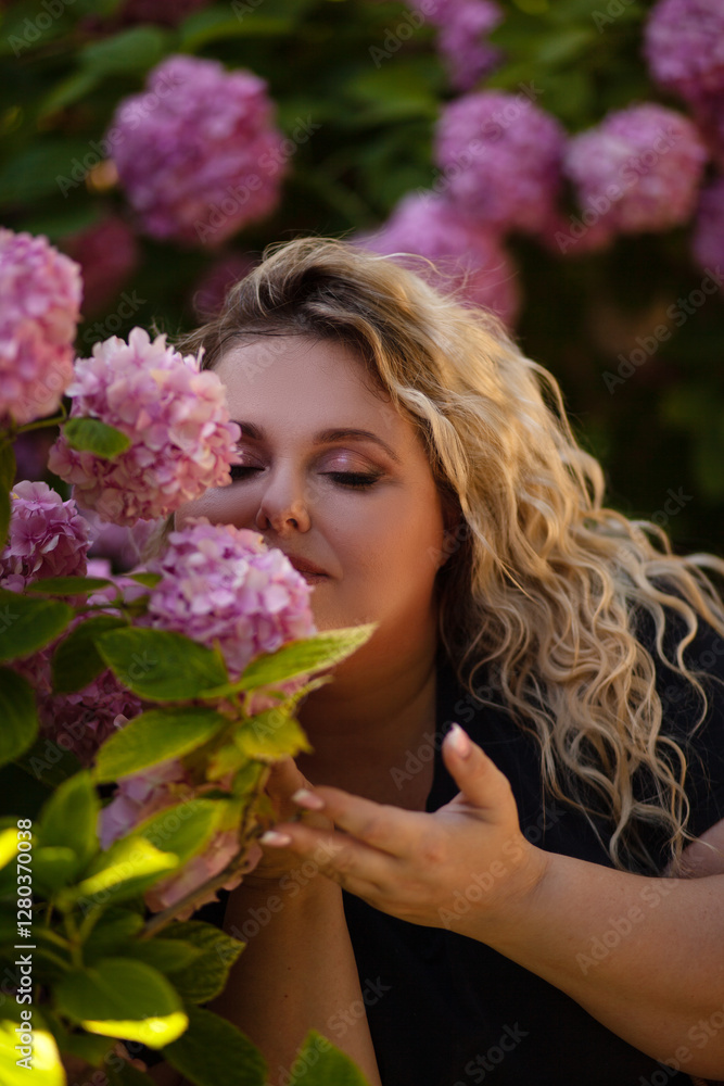 Portrait of a beautiful fat girl in a blue dress, against the background of a wall of live growing pink gartenias. The plus size woman enjoys the beauty of flowers Portrait of a beautiful fat girl in a blue dress, against the background of a wall of live growing pink gartenias. The plus size woman enjoys the beauty of flowers
