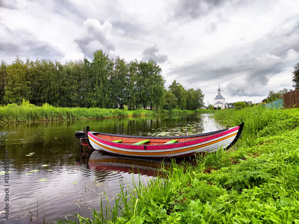 Colorful boat rests peacefully on a calm river surrounded by lush greenery and overcast skies Colorful boat rests peacefully on a calm river surrounded by lush greenery and overcast skies