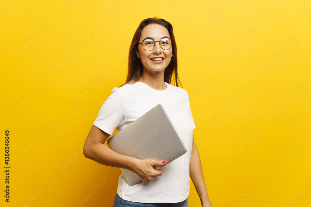 Smiling woman holding laptop poses against vibrant yellow background in bright indoor setting Smiling woman holding laptop poses against vibrant yellow background in bright indoor setting