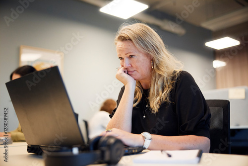 Thoughtful businesswoman hand on chin working on laptop at desk in office