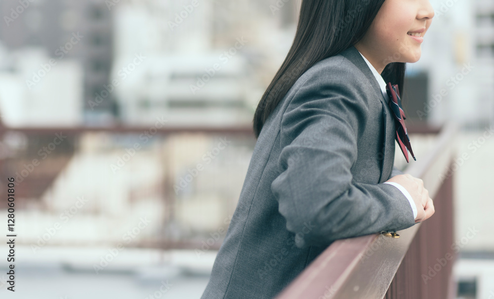 Fototapeta premium 制服を着た日本人の女の子が屋上から街の景色を眺めている。