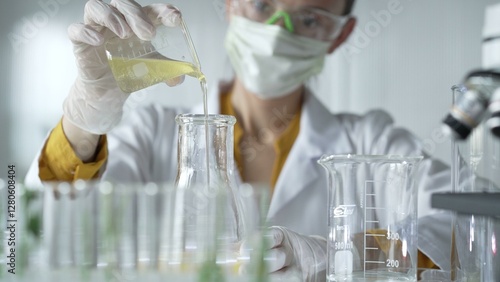Female scientist researcher wearing a lab coat, white gloves, and mask, is pouring a yellow oily liquid from a beaker into an Erlenmeyer flask in laboratory, portrait. Science and medicine concepts