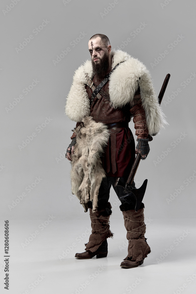 Fototapeta premium Full-length portrait of determined man, medieval Viking dressed in leather armor and fur cloak, standing with axe against grey studio background. Concept of historical portrait, Nordic mythology